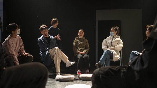 一人站在围坐的人群之间，手部动作可见
