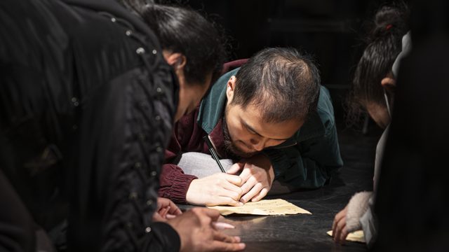 一人俯身用笔在纸上书写，旁有人围拢