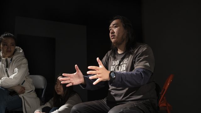 一人坐姿双手展开，旁人近处观看